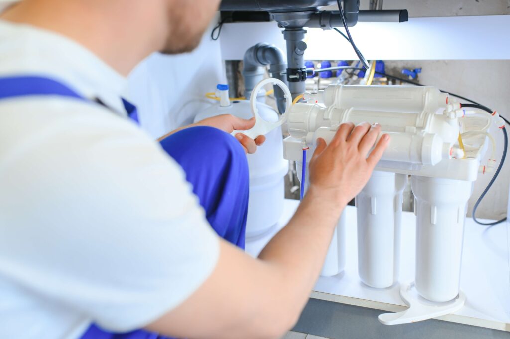 A blurry plumber is adjusting a reverse osmosis water filtration system under a kitchen sink in Lake County, FL, highlighting the system's white filter housings and pipes.