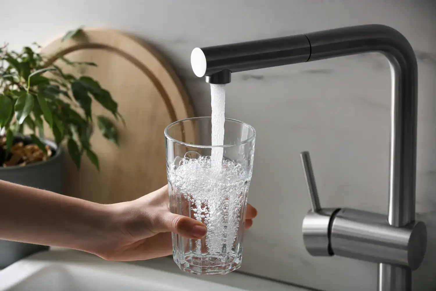 A close-up of a hand filling a clear glass with water from a modern stainless steel faucet in a kitchen in Lake County, FL, with a potted plant and wooden cutting board in the background.
