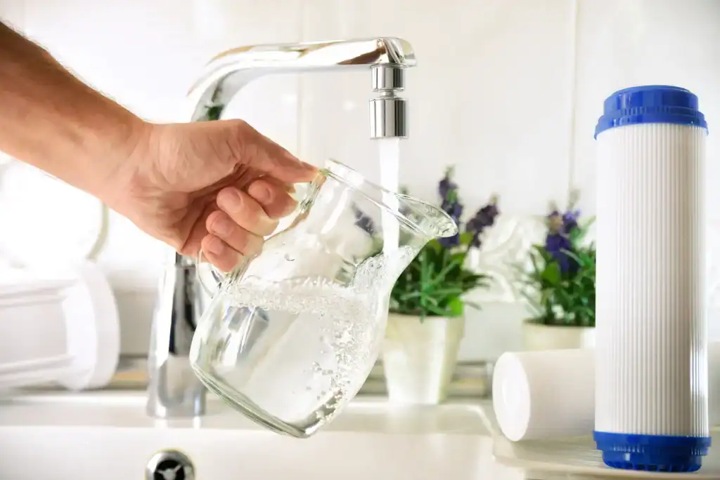 A hand holds a glass pitcher under a modern faucet, filling it with clear water. Two clean, white filter cartridges are visible on the counter to the right, emphasizing the purity of the filtered water in Lake County, FL.
