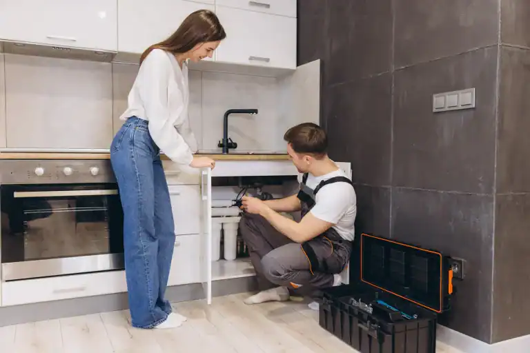 A plumber is installing or repairing a water filtration system under a kitchen sink in a home in Lake County, FL, while a woman observes.