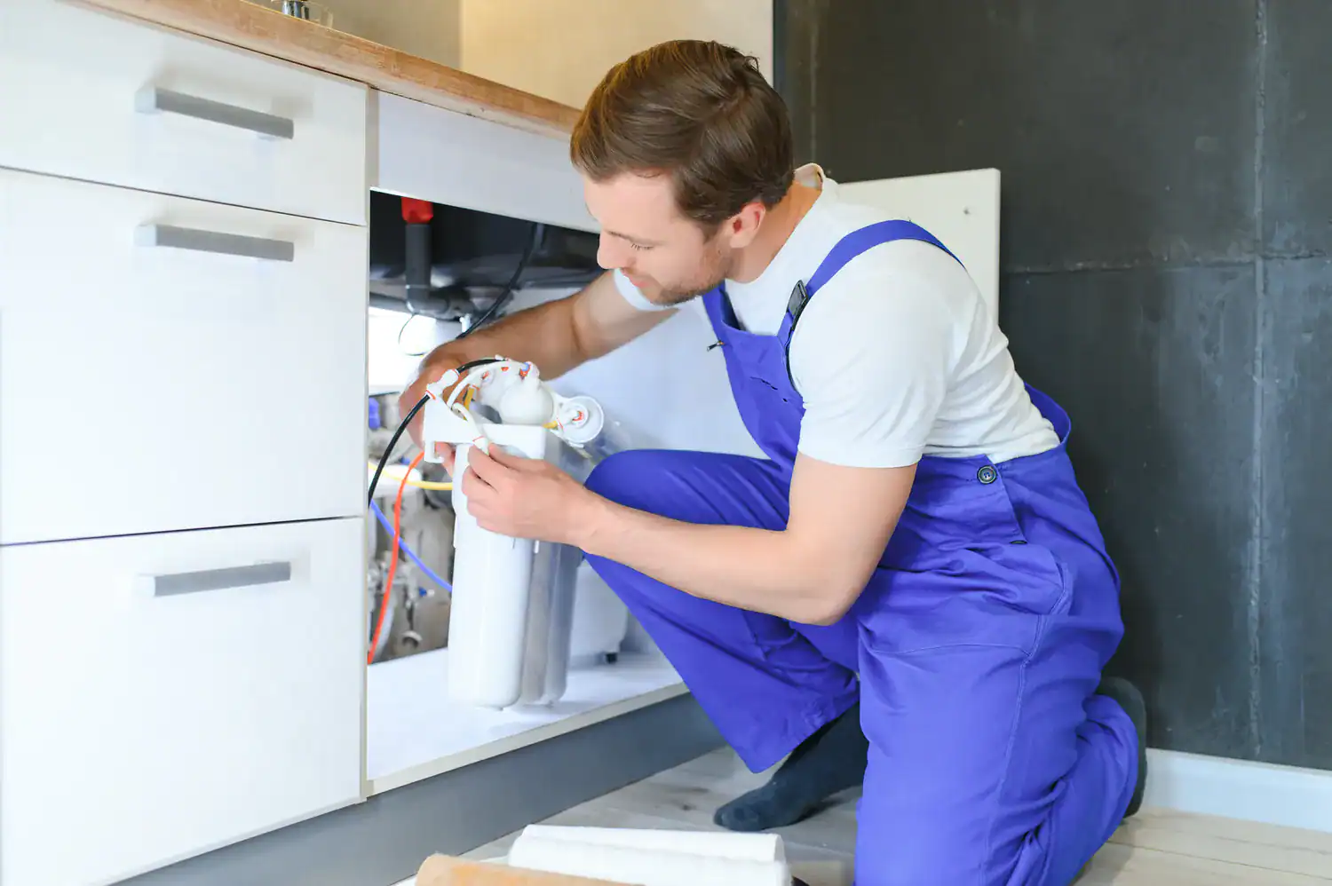 A professional plumber in Lake County, FL, is kneeling and working on a water filtration system installed under a kitchen sink.