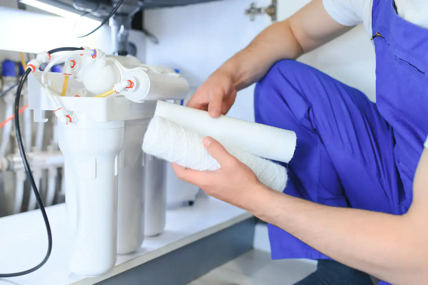 A plumber in blue overalls is holding two new filter cartridges, preparing to install them into a reverse osmosis water filtration system under a sink in Lake County, FL.