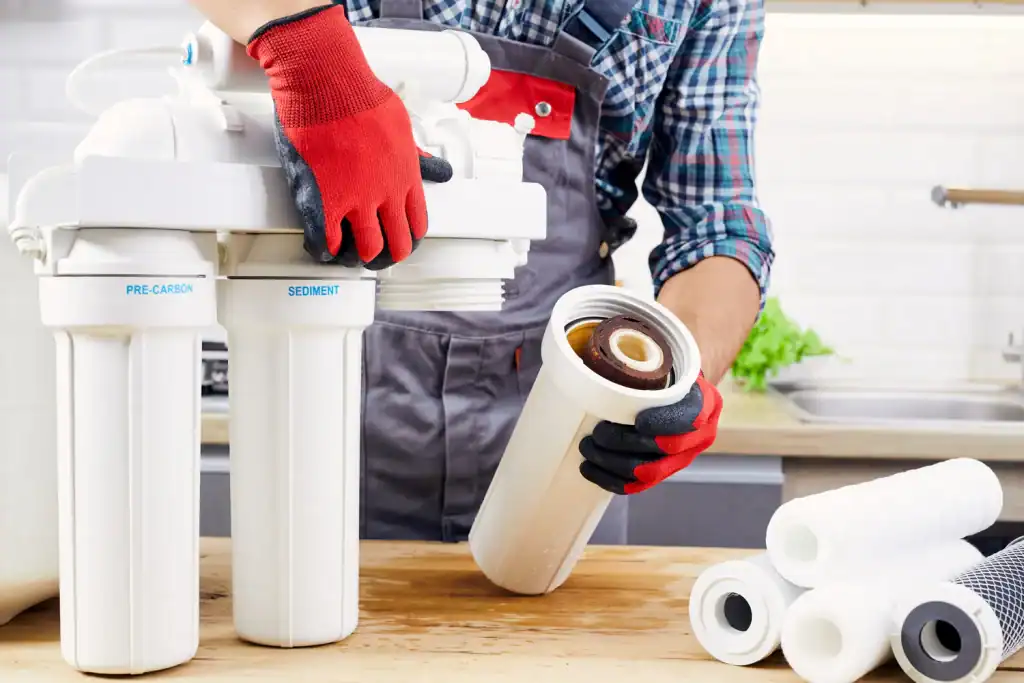 A service technician wearing red and black gloves changes a filter cartridge in a multi-stage water filtration system, with new filter cartridges stacked nearby on a wooden table in Lake County, FL.