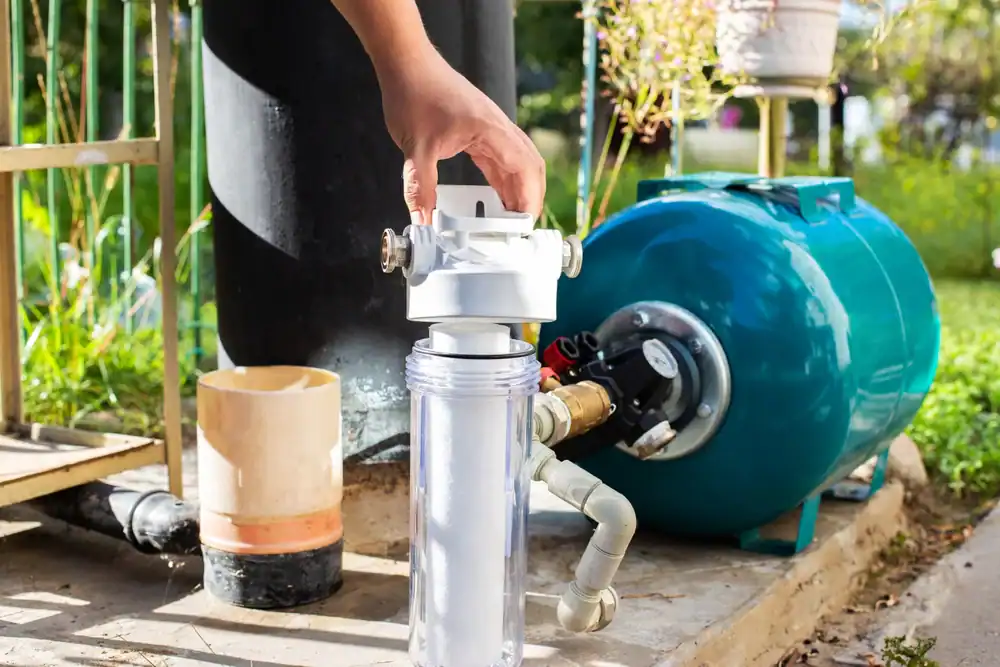 A hand adjusts a water filtration system connected to a blue pressure tank in an outdoor setup in Lake County, FL, surrounded by pipes, fittings, and greenery.