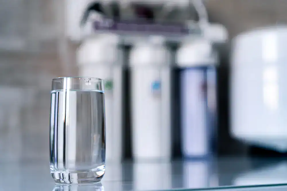 A clear glass of water sits on a reflective surface with a blurred water filtration system in the background, suggesting purified water in Lake County, FL.