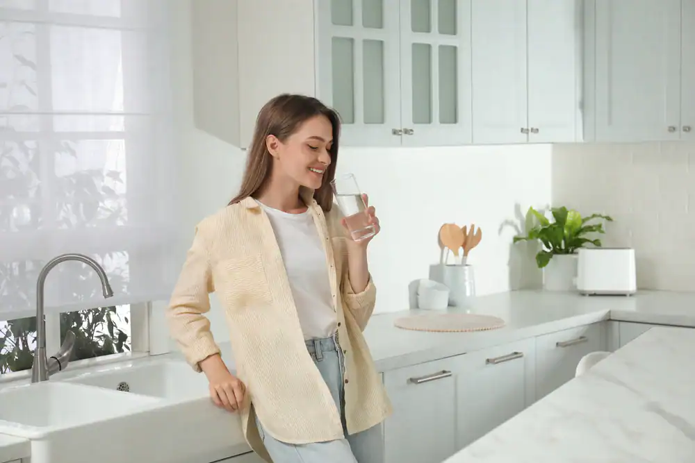 A happy woman enjoys a glass of clean, filtered water while standing in a bright kitchen in Lake County, FL, highlighting the benefits of home water purification.