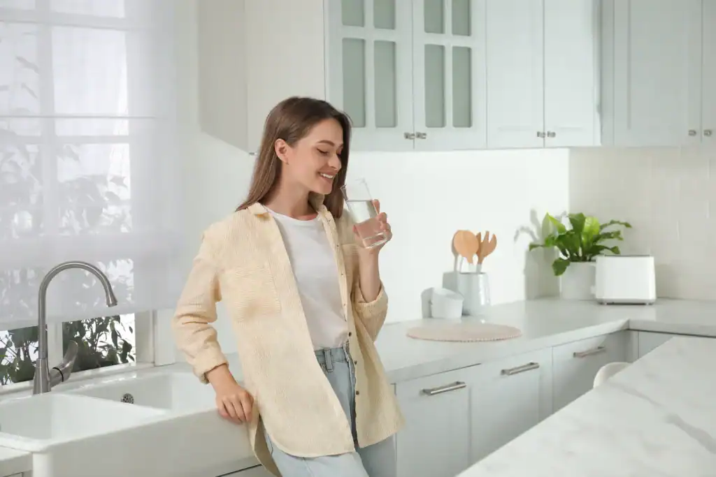 A happy young woman in Lake County, FL, leans against a kitchen counter and smiles while holding a glass of clean, filtered drinking water.