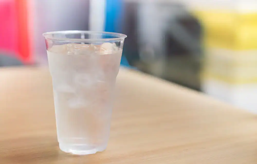 A clear plastic cup filled with ice water sits on a light wooden table, highlighting the purity provided by FL Water Filtration Systems Lake County, with a blurred colorful background.