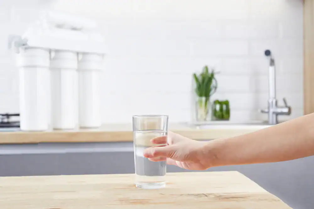 A hand reaches for a clear glass of water on a wooden kitchen counter, with a Water Filtration Systems Lake County, FL setup and sink visible in the background.