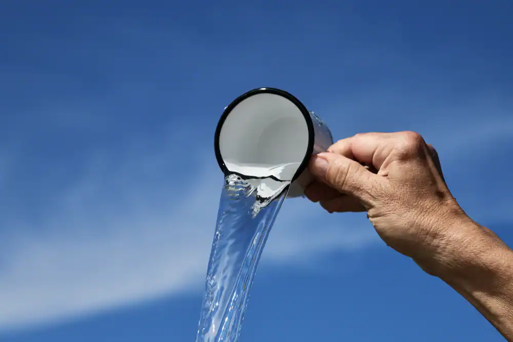 A hand tilts a white enamel cup with a chipped rim, pouring a stream of clear water&mdash;thanks to Water Filtration Systems Lake County, FL&mdash;against a bright blue sky background.