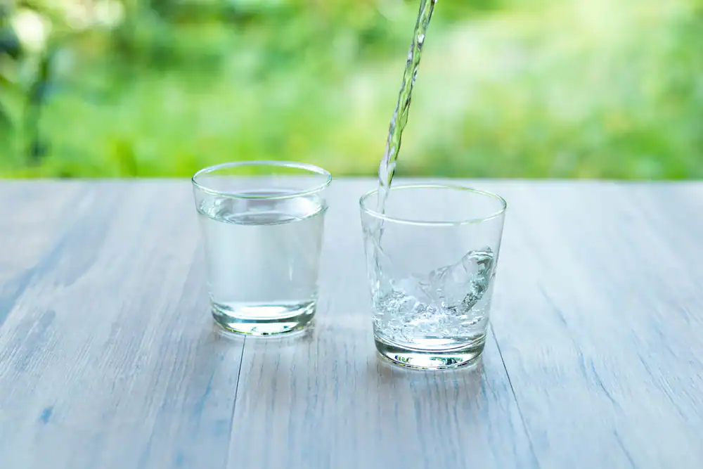 Two clear glasses sit on a light wooden surface; one is filled with water, while water from Water Filtration Systems Lake County, FL is being poured into the other. The background is blurred greenery.