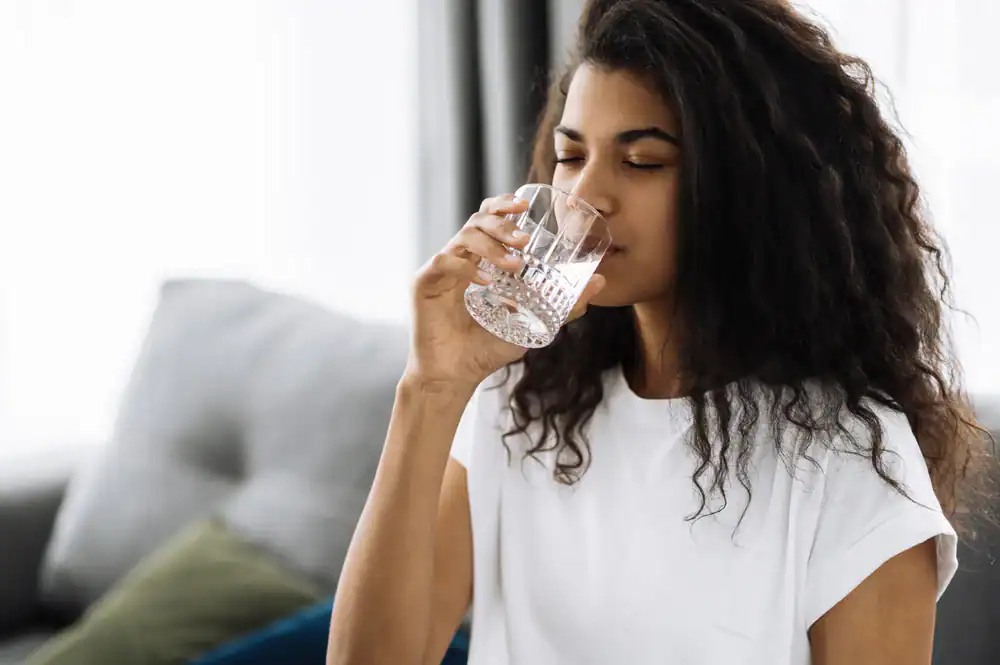 A young woman with long, curly hair wearing a white t-shirt is drinking a glass of water indoors. She has her eyes closed and appears relaxed, enjoying the refreshing taste from Water Filtration Systems Lake County, FL. The background is softly blurred.