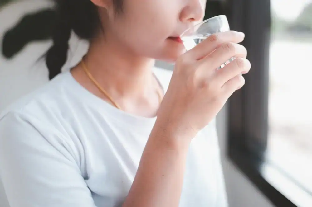 A person with a braid, wearing a white shirt and a gold necklace, enjoys a glass of water near a window in FL, appreciating the benefits of Water Filtration Systems Lake County under soft natural light.