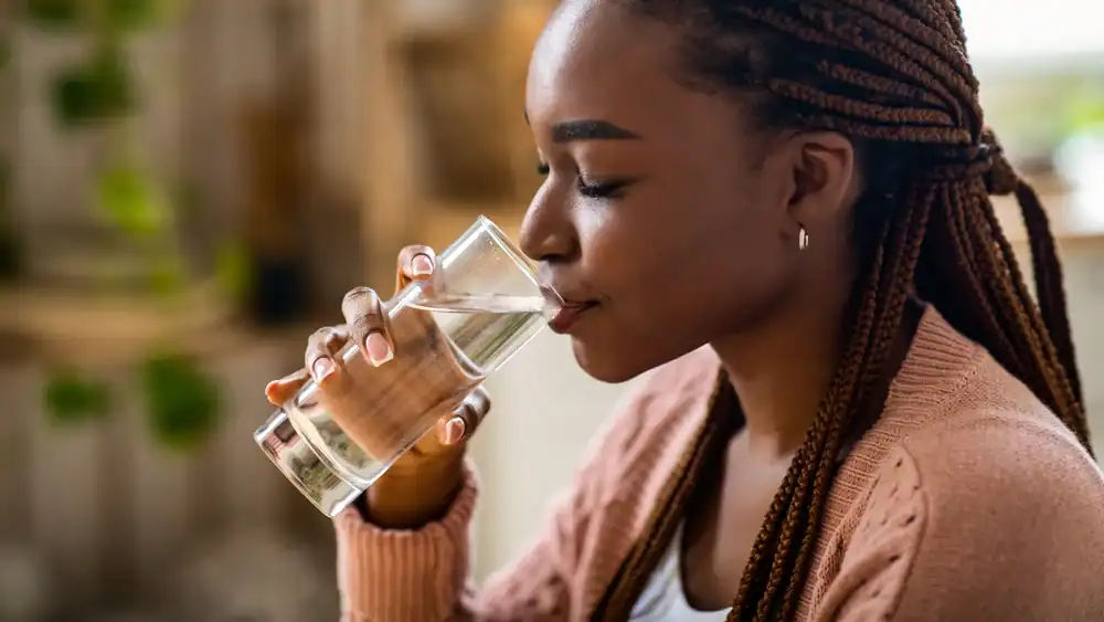 A young woman with braided hair is sitting indoors and drinking a glass of water. She is wearing a light pink cardigan and appears relaxed, enjoying a sip thanks to Water Filtration Systems Lake County, FL.