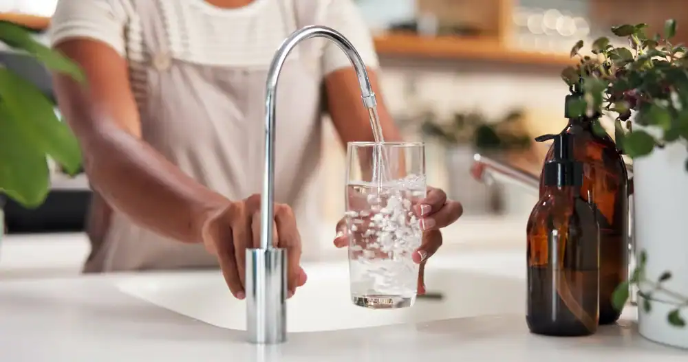 A person in FL holds a glass under a modern kitchen faucet, filling it with water. Plants and a brown bottle sit on the counter next to the sink, highlighting the benefits of Water Filtration Systems Lake County residents rely on.