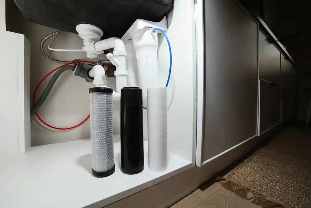 Three water filter cartridges, part of advanced Water Filtration Systems Lake County, FL, are placed in front of plumbing pipes under a kitchen sink, surrounded by white cabinets, a section of countertop, and a brown rug on the floor.