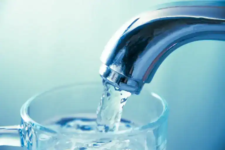 A close-up of water flowing from a shiny metal faucet into a clear glass, with a light blue background, highlights the benefits of Water Filtration Systems Lake County, FL residents can trust for fresh and clean drinking water.