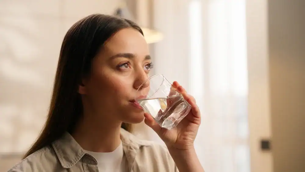 A woman with long dark hair is indoors, holding a glass of water and drinking from it&mdash;enjoying the fresh taste made possible by Water Filtration Systems Lake County, FL. She is looking slightly upward, wearing a light-colored shirt in a softly lit room.